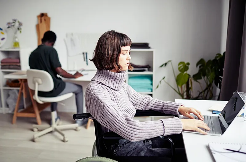 Kobieta na wózku inwalidzkim pracująca przy laptopie w biurowym otoczeniu, ilustrująca aktywizację zawodową osób niepełnosprawnych i procesy PFRON